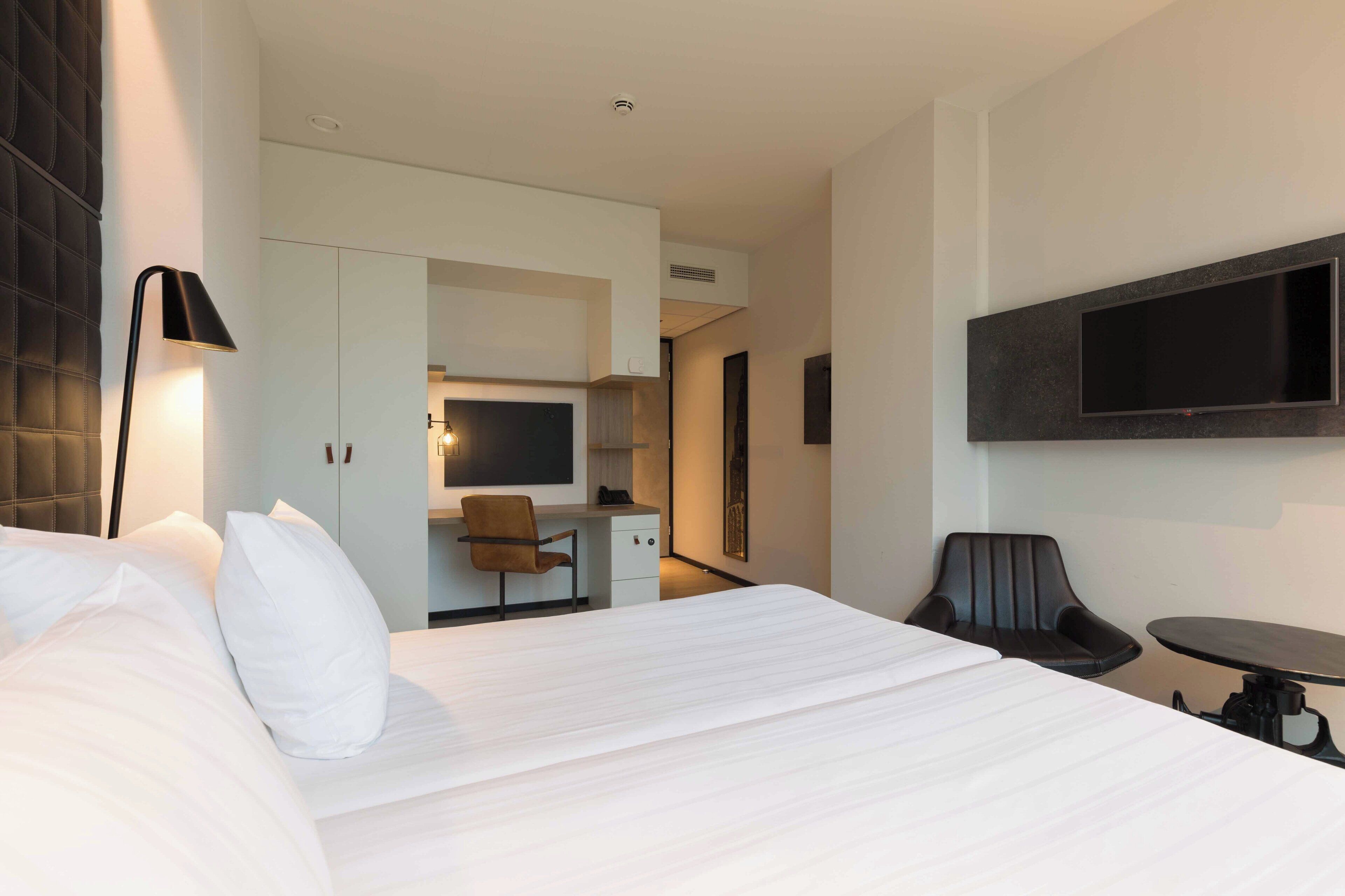 A modern hotel room with a large bed, a desk, a chair, a TV mounted on the wall, and a small table.