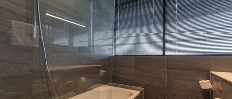 Un baño moderno con bañera con mampara de vidrio, inodoro y ventana con persianas.