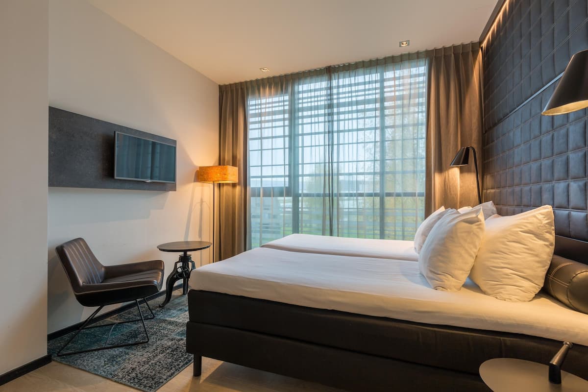 Habitación de hotel moderna con una cama grande, una silla, una mesita pequeña, un televisor montado en la pared y un ventanal con cortinas.