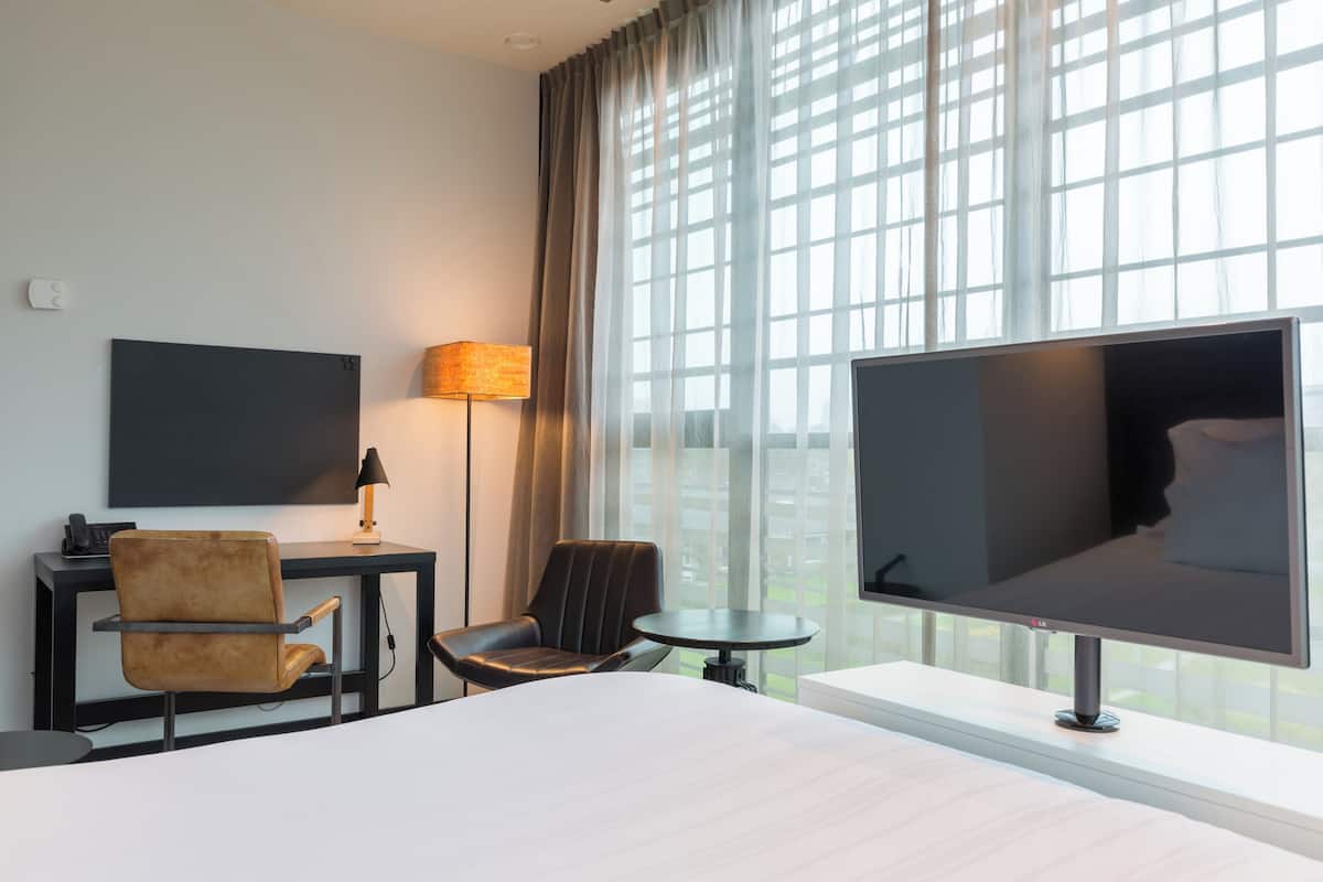 Habitación de hotel con cama, escritorio con lámpara, silla, televisor y ventanas amplias.