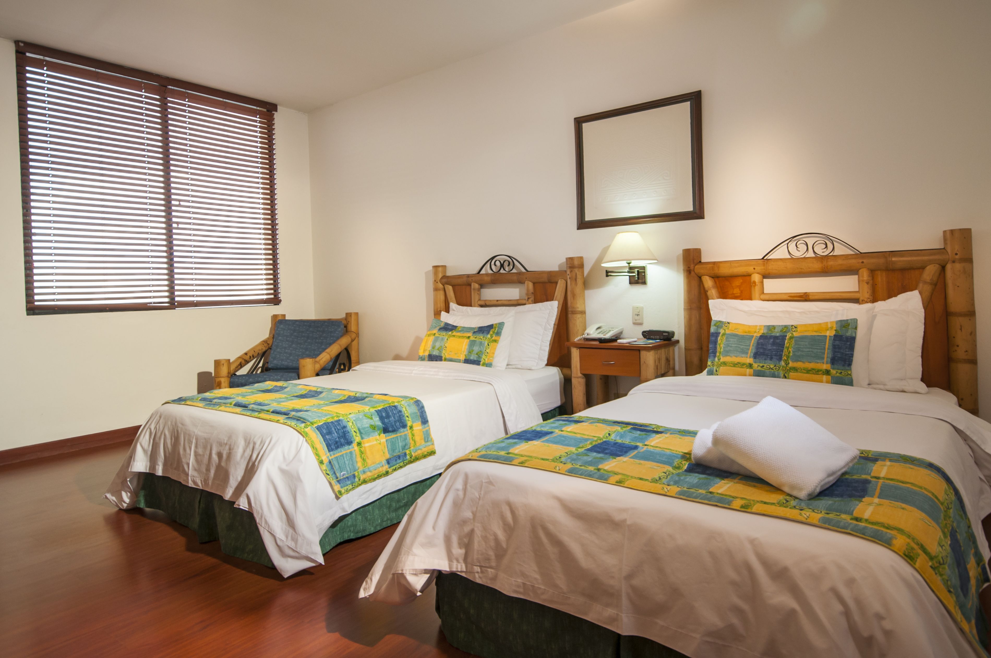 A hotel room with two beds, wooden headboards, a nightstand with a lamp, and a window with blinds.
