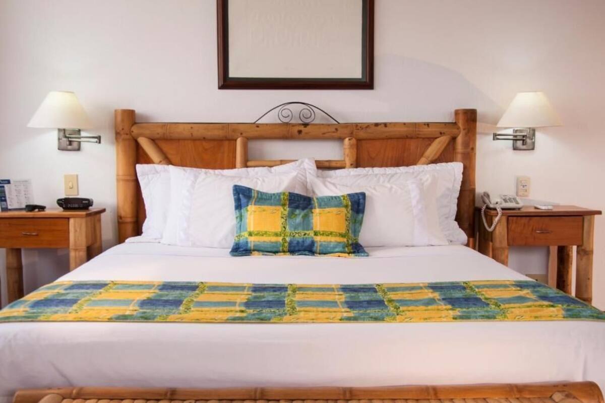 A bed with a wooden headboard, two bedside tables, a wall-mounted lamp, a framed picture, and a patterned bedspread.