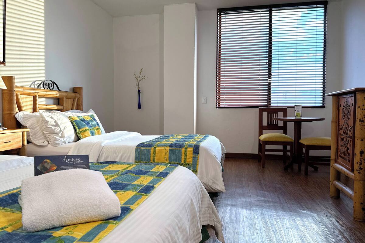 A hotel room with two beds, a wooden nightstand, a desk with a chair, and a window with blinds.