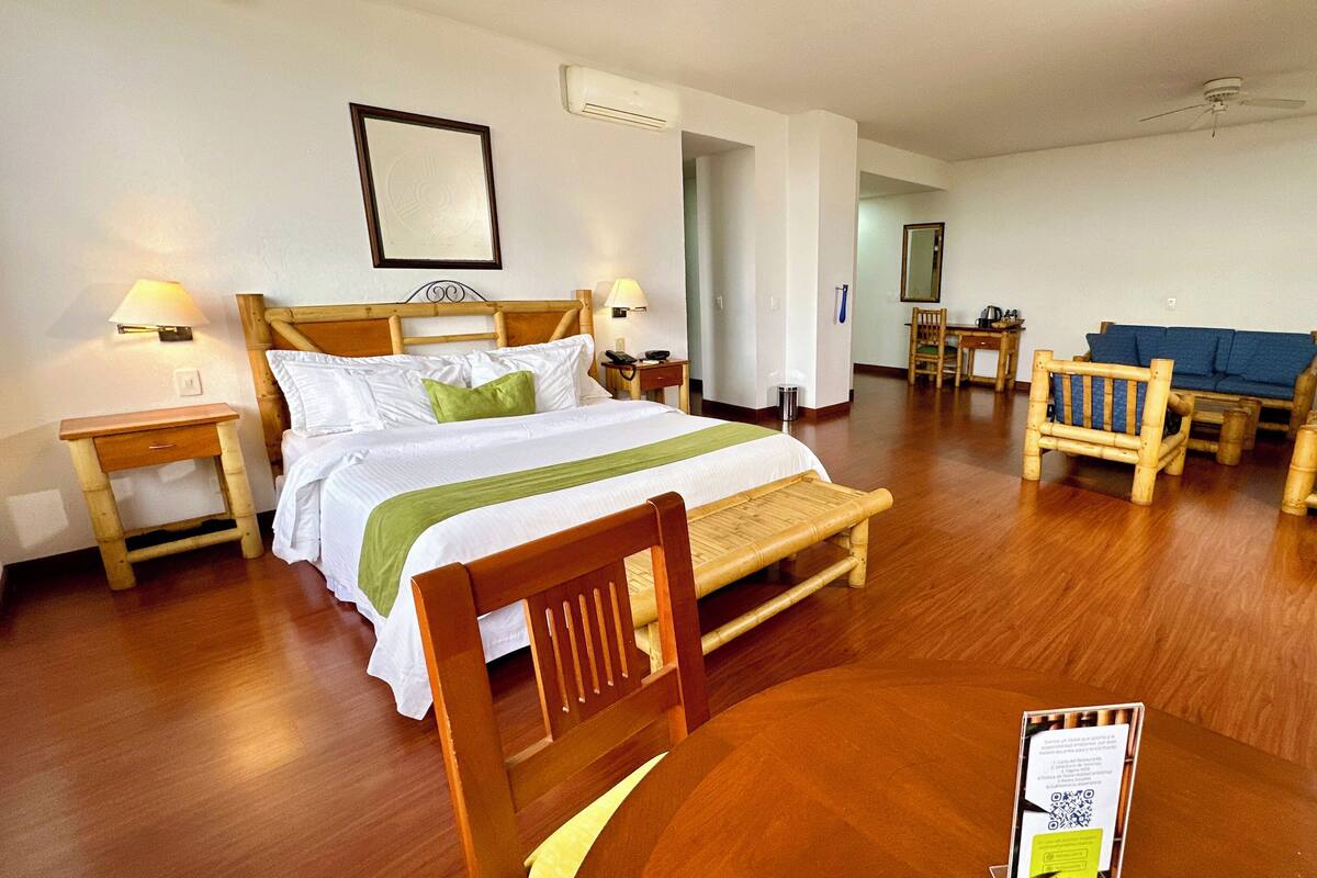 A hotel room with a wooden bed, a wooden chair, a small table, and a bedside table with a lamp.