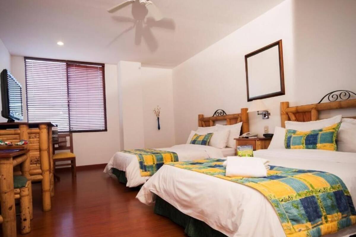 A hotel room with two beds, a wooden desk, a chair, a ceiling fan, and a window with blinds.