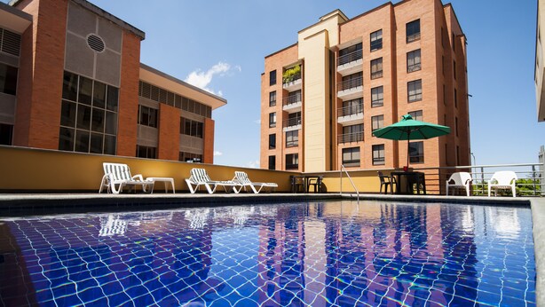 Une piscine avec un sol carrelé et des chaises longues. On aperçoit deux bâtiments à l’arrière-plan, l’un avec une façade en briques et l’autre avec une façade beige.