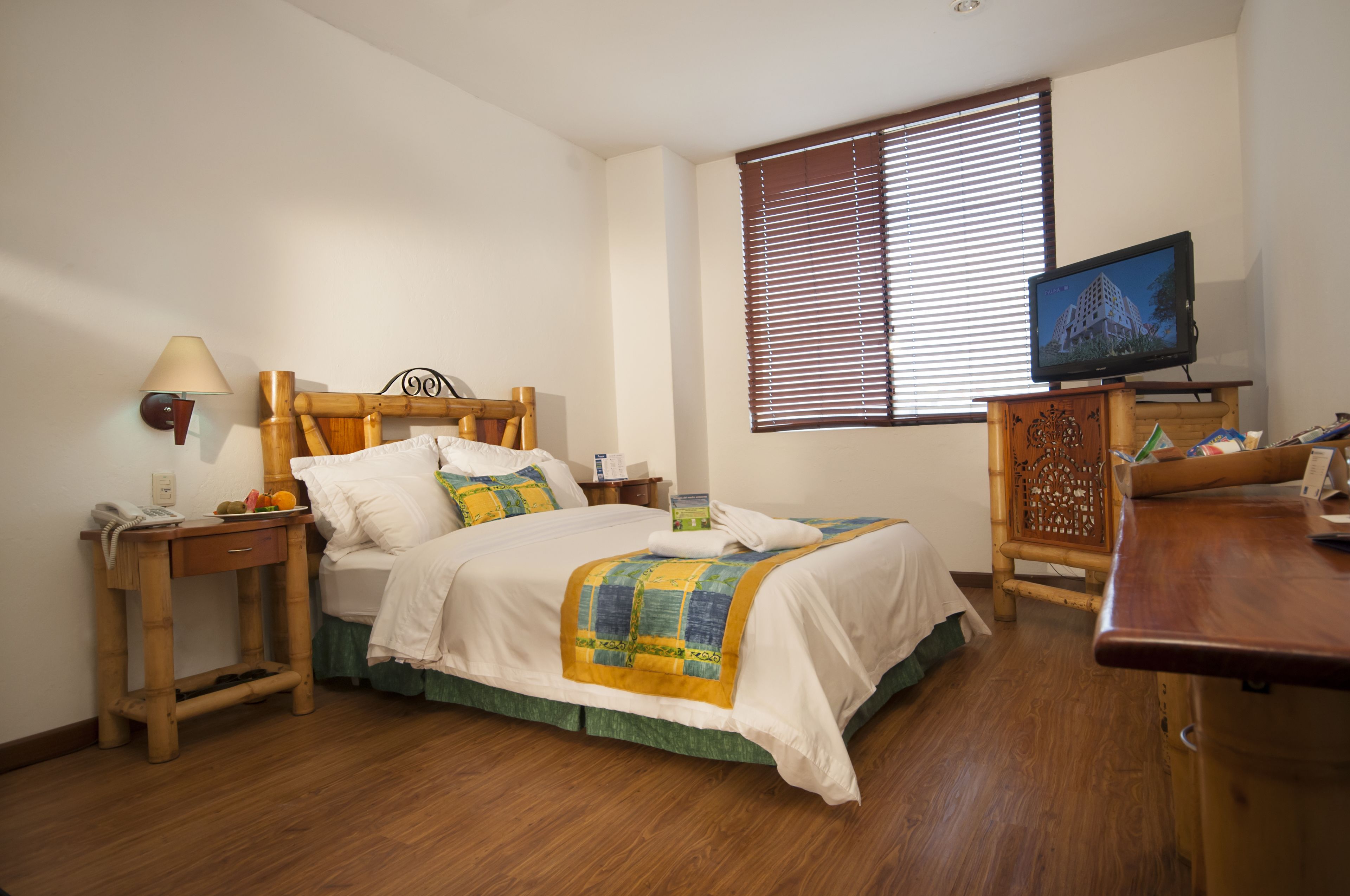 Un dormitorio ordenado con cama, mesitas de noche, televisor y ventana con persianas.