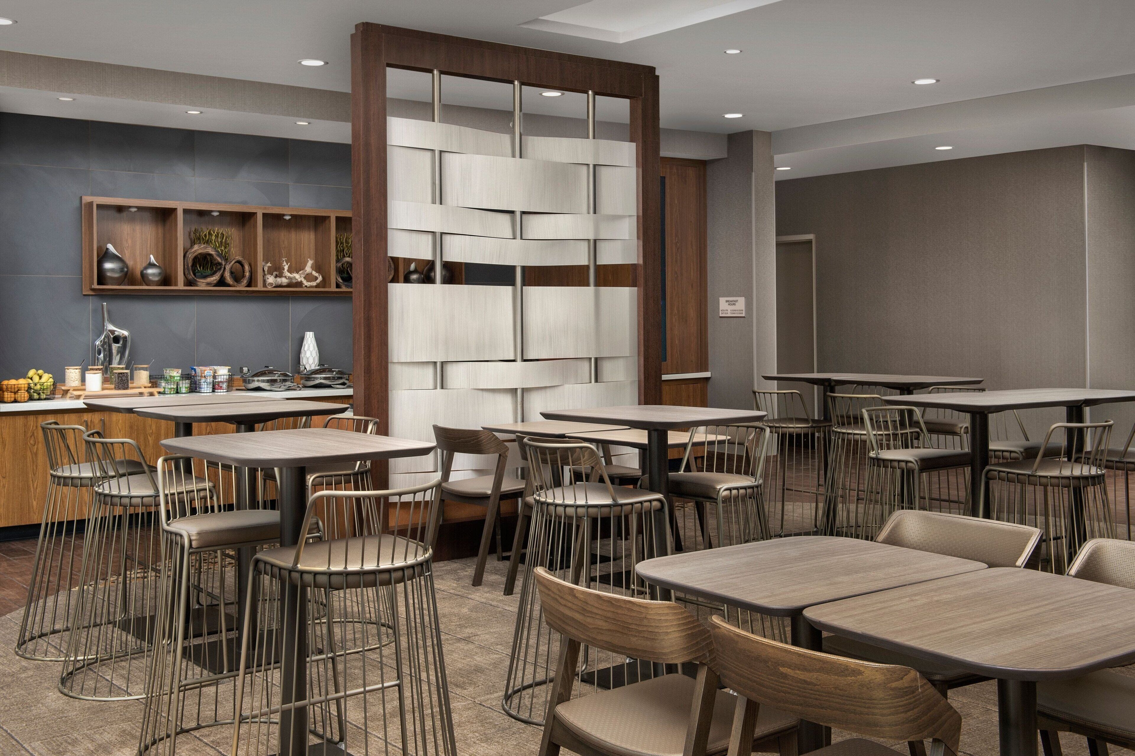 Un comedor moderno con varias mesas y sillas, un separador de madera con estantes y una zona de cocina con distintos objetos a la vista.