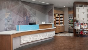 Un área de recepción con diseño moderno, que incluye un mostrador con cubierta azul y base de madera, una pared con diseño y una exhibición de aperitivos y bebidas.
