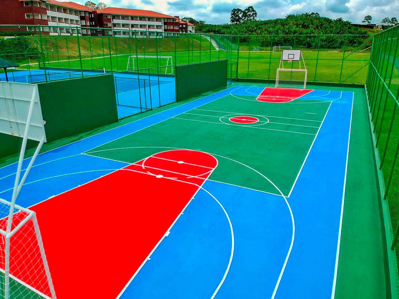 An outdoor basketball court with a red and blue playing surface, surrounded by a green fence.