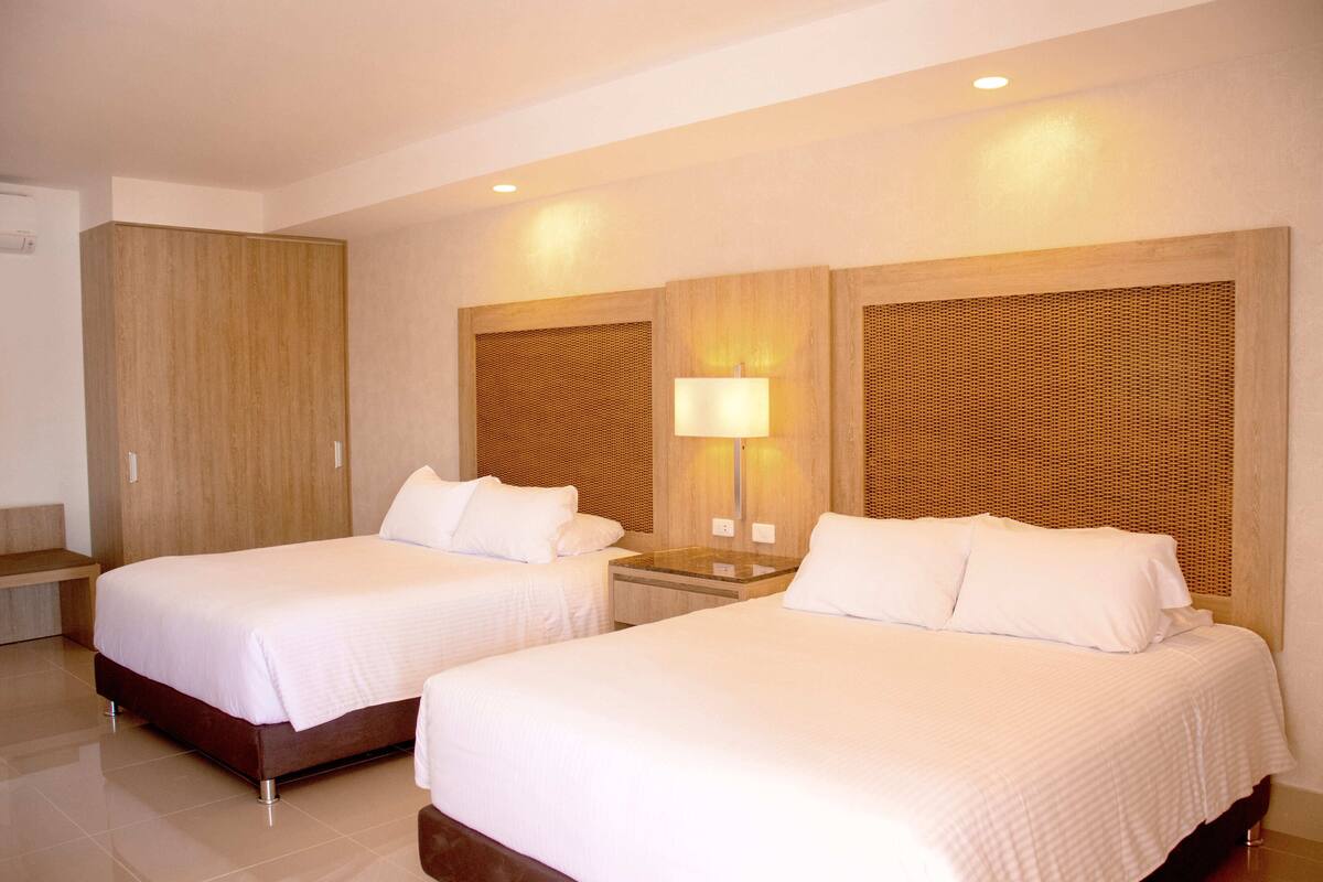 Una habitación de hotel con dos camas, un armario de madera, una mesita de noche con lámpara y un banco.