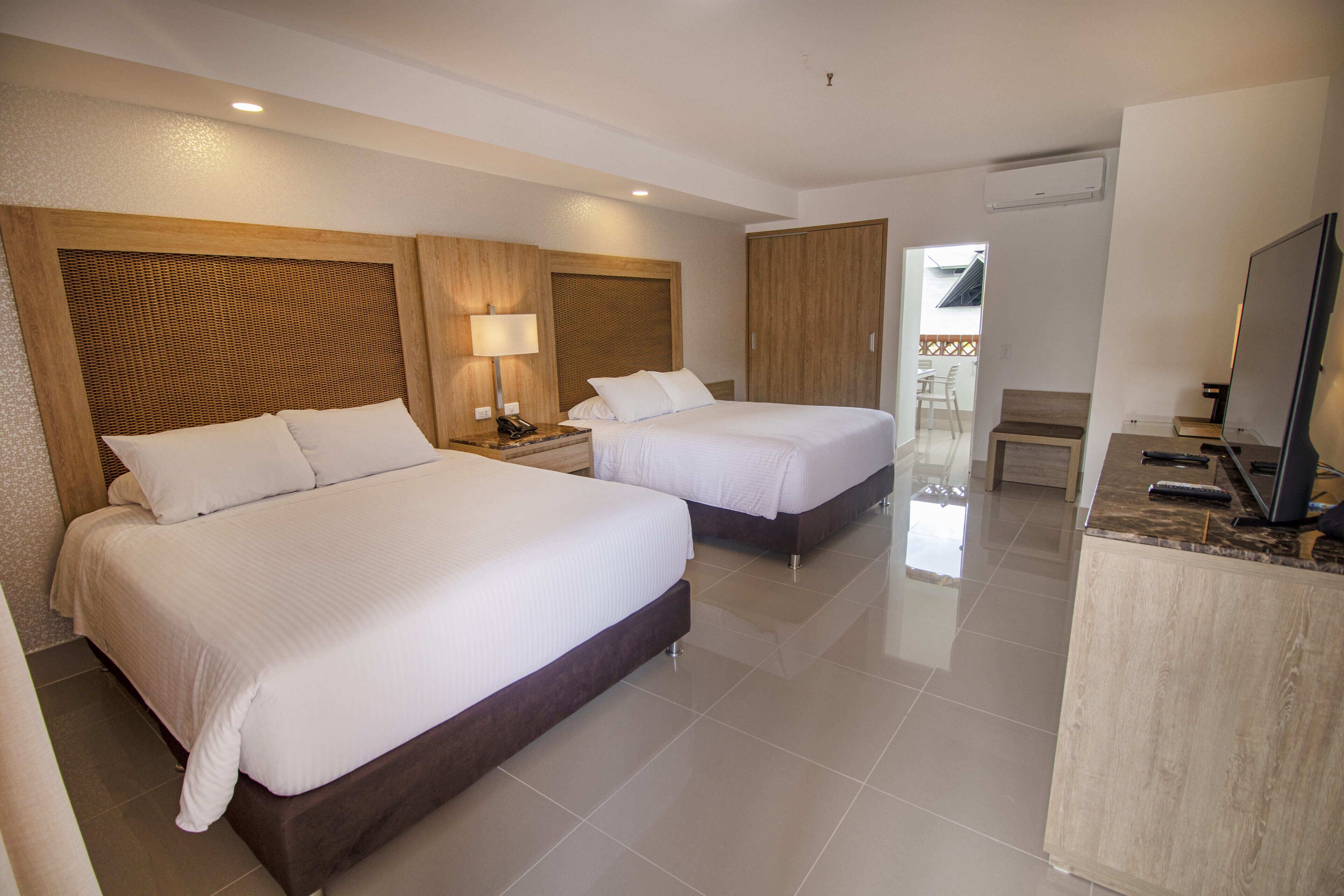 A hotel room with two beds, a desk with a TV, and a balcony visible through the door.