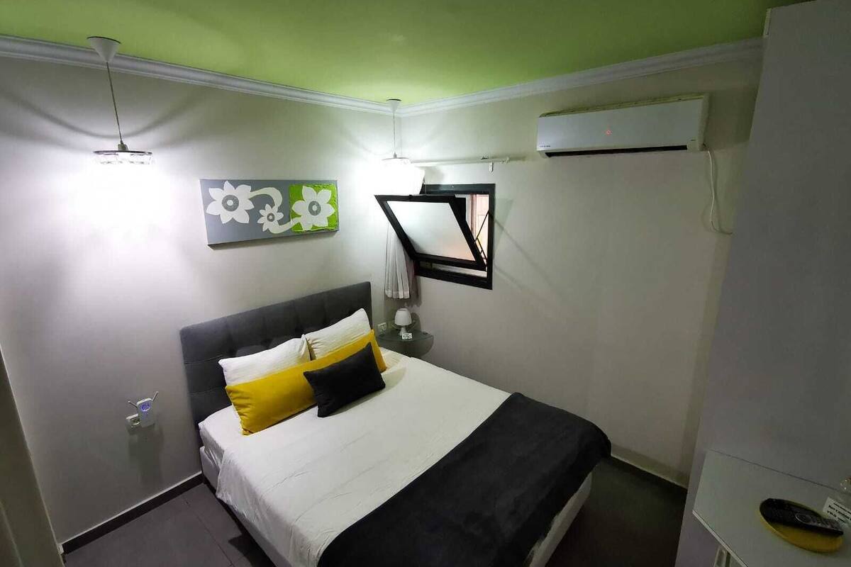 A bedroom with a bed, a nightstand, a wall-mounted air conditioner, and a window with a view.