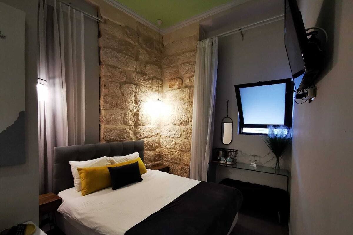 A bedroom with a stone wall, a bed with white and yellow bedding, a desk with a computer, and a TV mounted on the wall.