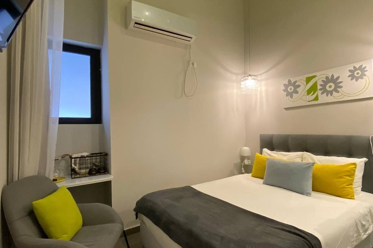 A bedroom with a bed, a chair, an air conditioning unit, a window, and a wall-mounted light fixture.