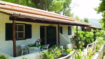 A building with a tiled roof, multiple windows with shutters, a covered porch area with outdoor seating, and lush greenery surrounding the structure.