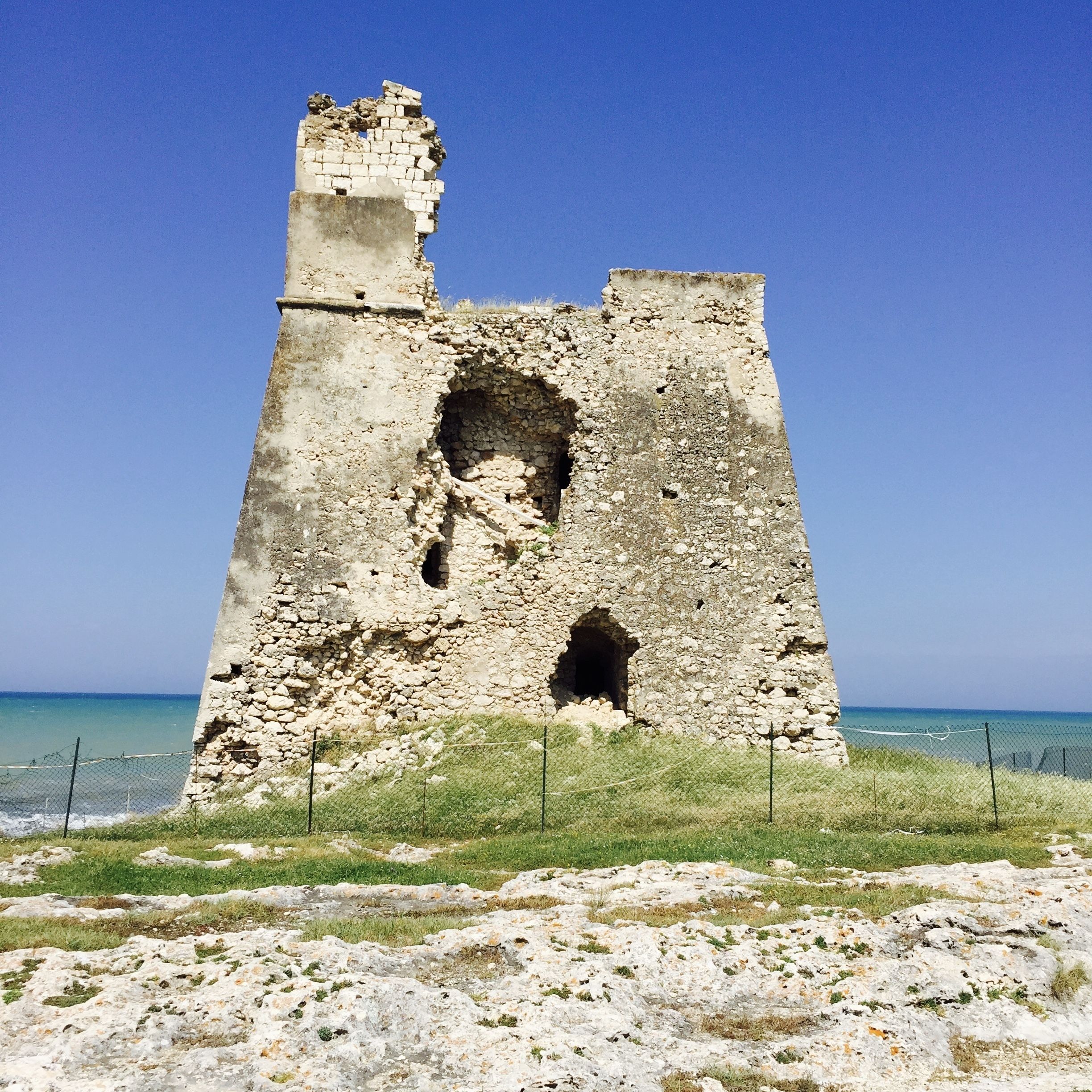 A large, ancient stone structure with a tower and arched openings, surrounded by a grassy area and a fence, overlooking a clear blue sea.