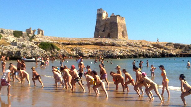 Una playa con gente en traje de baño, una estructura parecida a un castillo al fondo y un litoral rocoso.