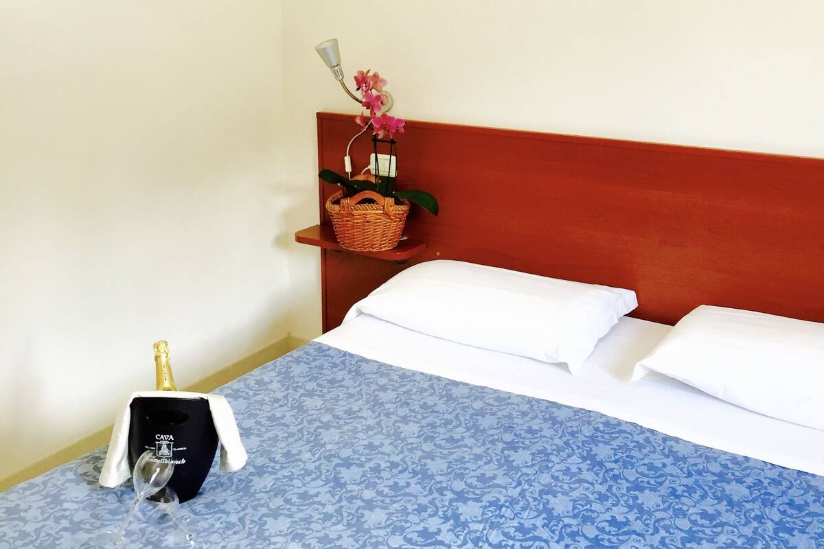 A hotel room with a bed, a nightstand, a bottle of champagne, a basket with flowers, and a bottle of water.