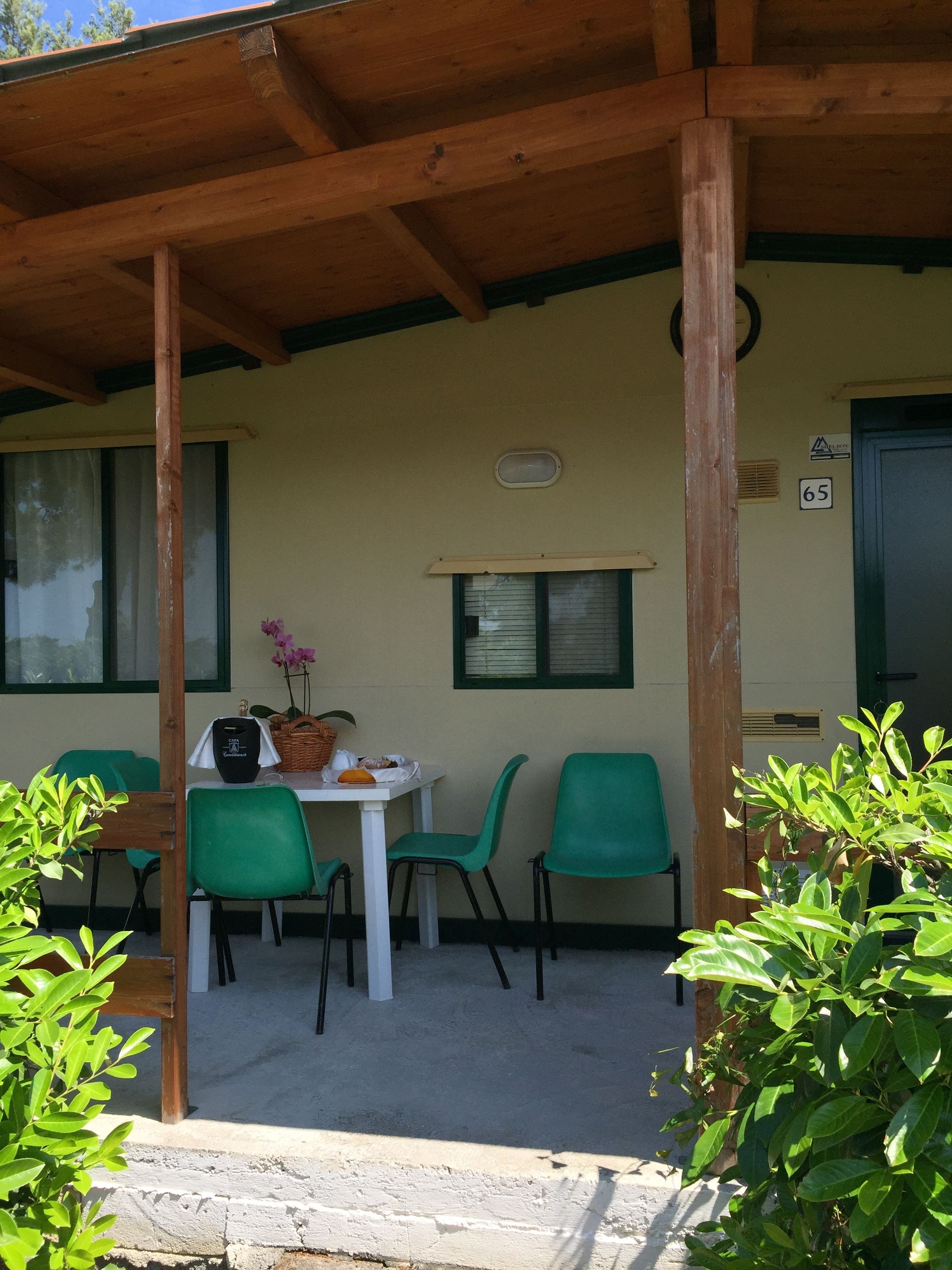 A covered outdoor seating area with a table and chairs, a window with blinds, and a door marked with the number 65.