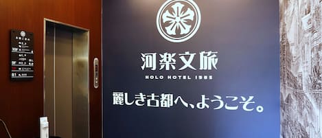 Un vestíbulo de hotel con una gran pared mostrando el nombre y el logotipo del hotel, un ascensor y una pequeña mesa con dispensador de agua.