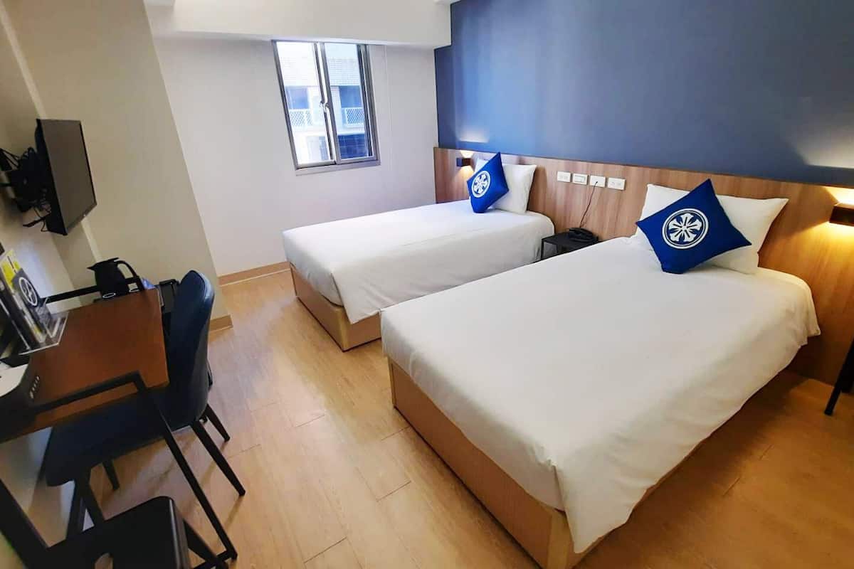 A hotel room with two beds, a desk, a chair, a TV, and a window.