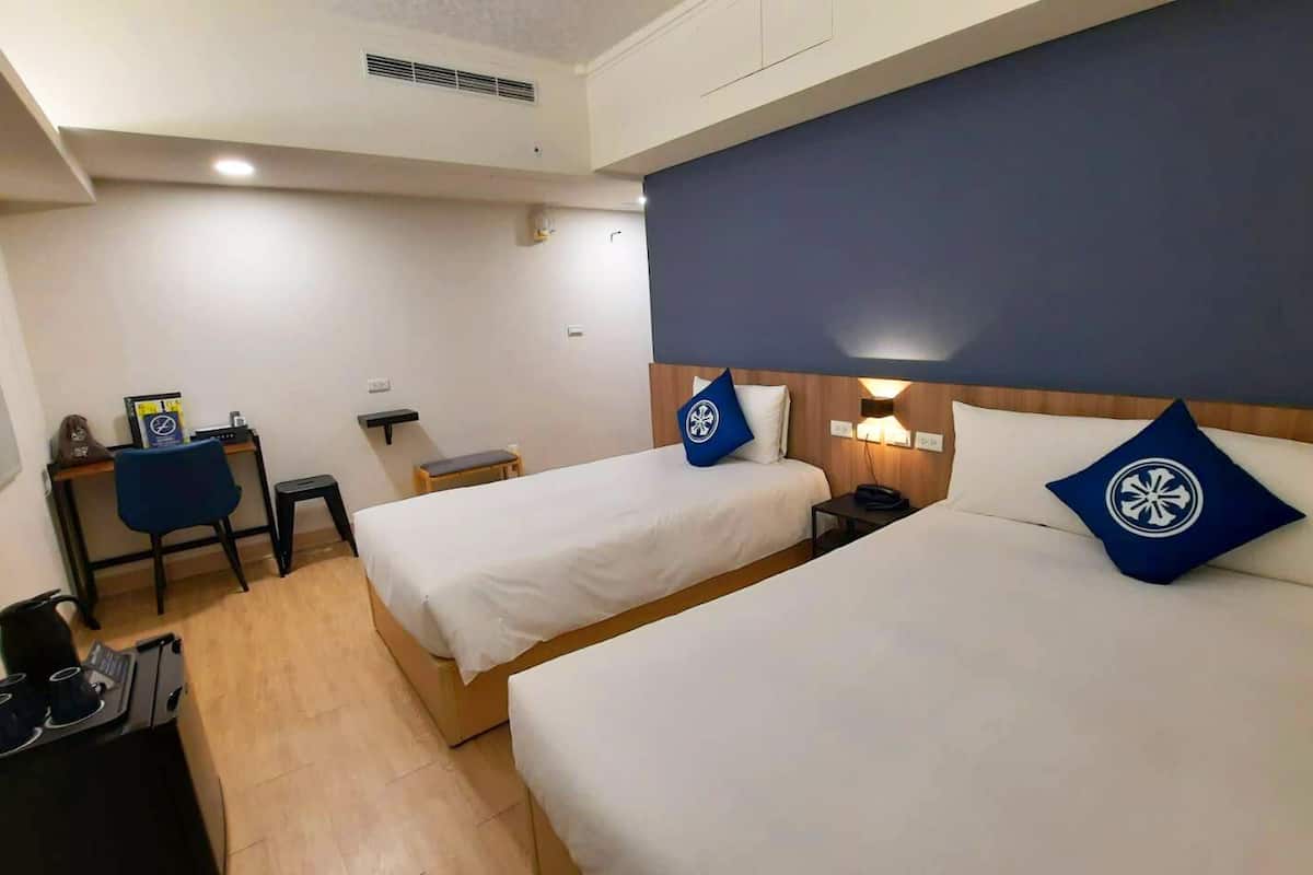 A hotel room with two beds, a desk, a chair, and a small table with cups and a kettle.