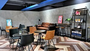 Un área de descanso moderna con un sofá de cuero marrón, sillas negras y anaranjadas, una mesita redonda y una estantería con objetos decorativos.