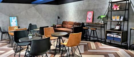 Un área de descanso moderna con un sofá de cuero marrón, sillas negras y anaranjadas, una mesita redonda y una estantería con objetos decorativos.