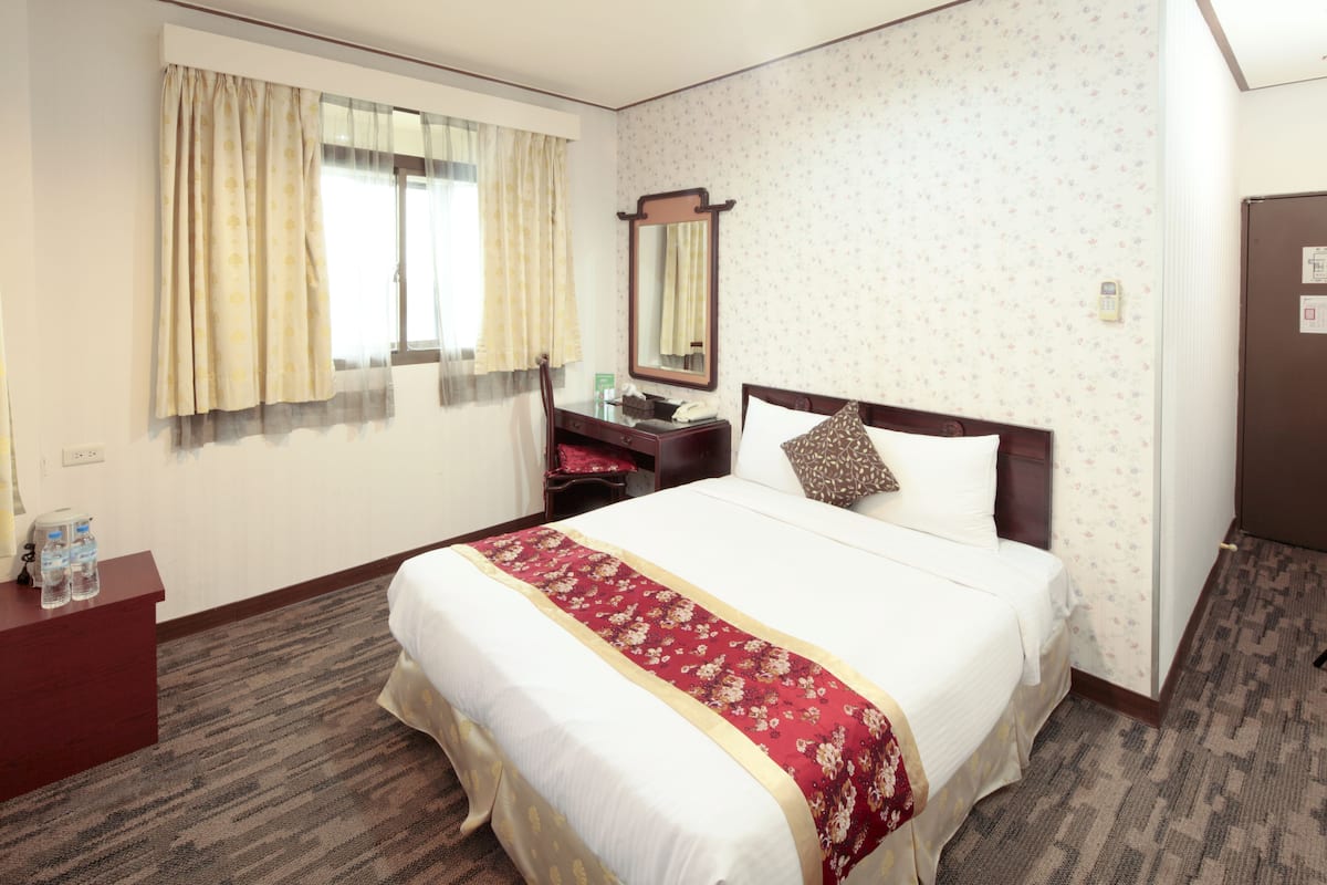 A hotel room with a large bed, a desk with a telephone, a mirror, and a window with curtains.