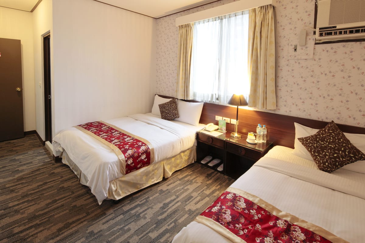 A hotel room with two beds, a nightstand, a lamp, and a window with curtains.