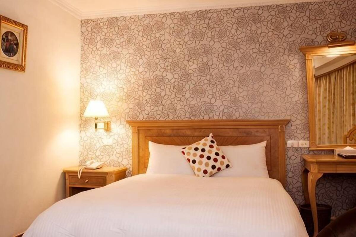 A hotel room with a patterned wallpaper, a wooden headboard, a bedside lamp, a nightstand, and a mirror.