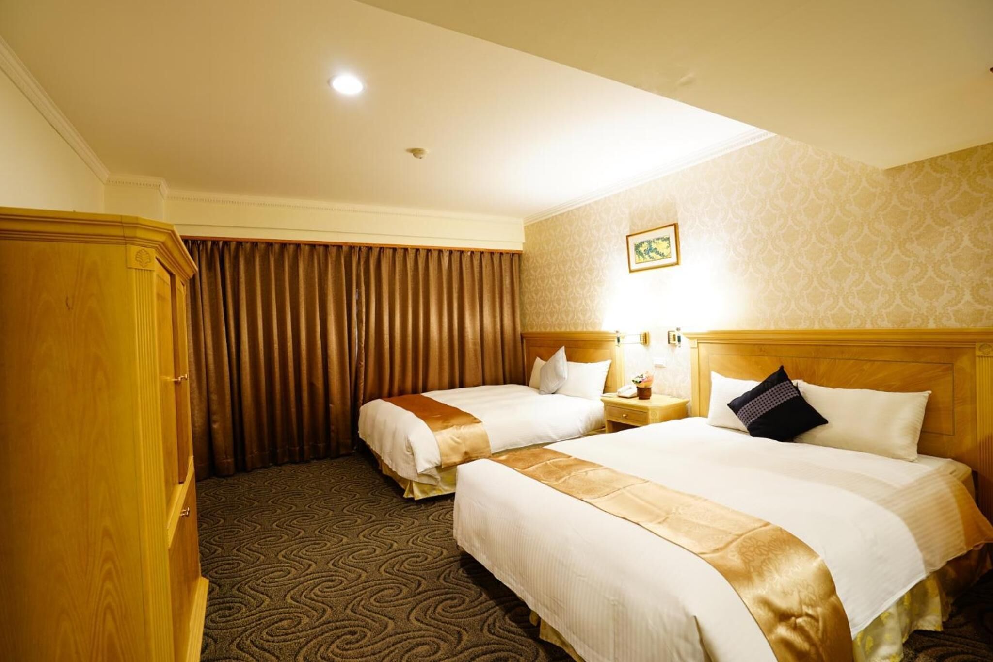 A hotel room with two beds, a wooden wardrobe, a patterned carpet, and a wall-mounted picture.