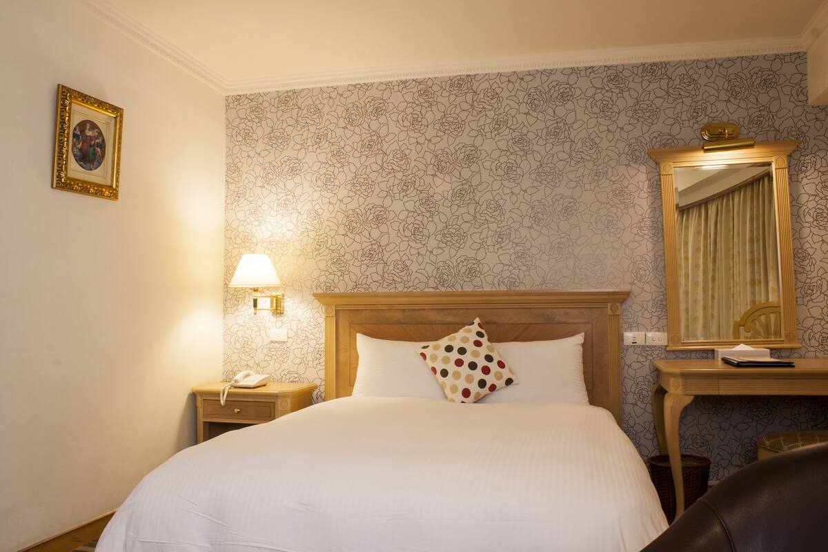 A hotel room with a large bed, a nightstand with a lamp, a telephone, a mirror, and a desk with a lamp.