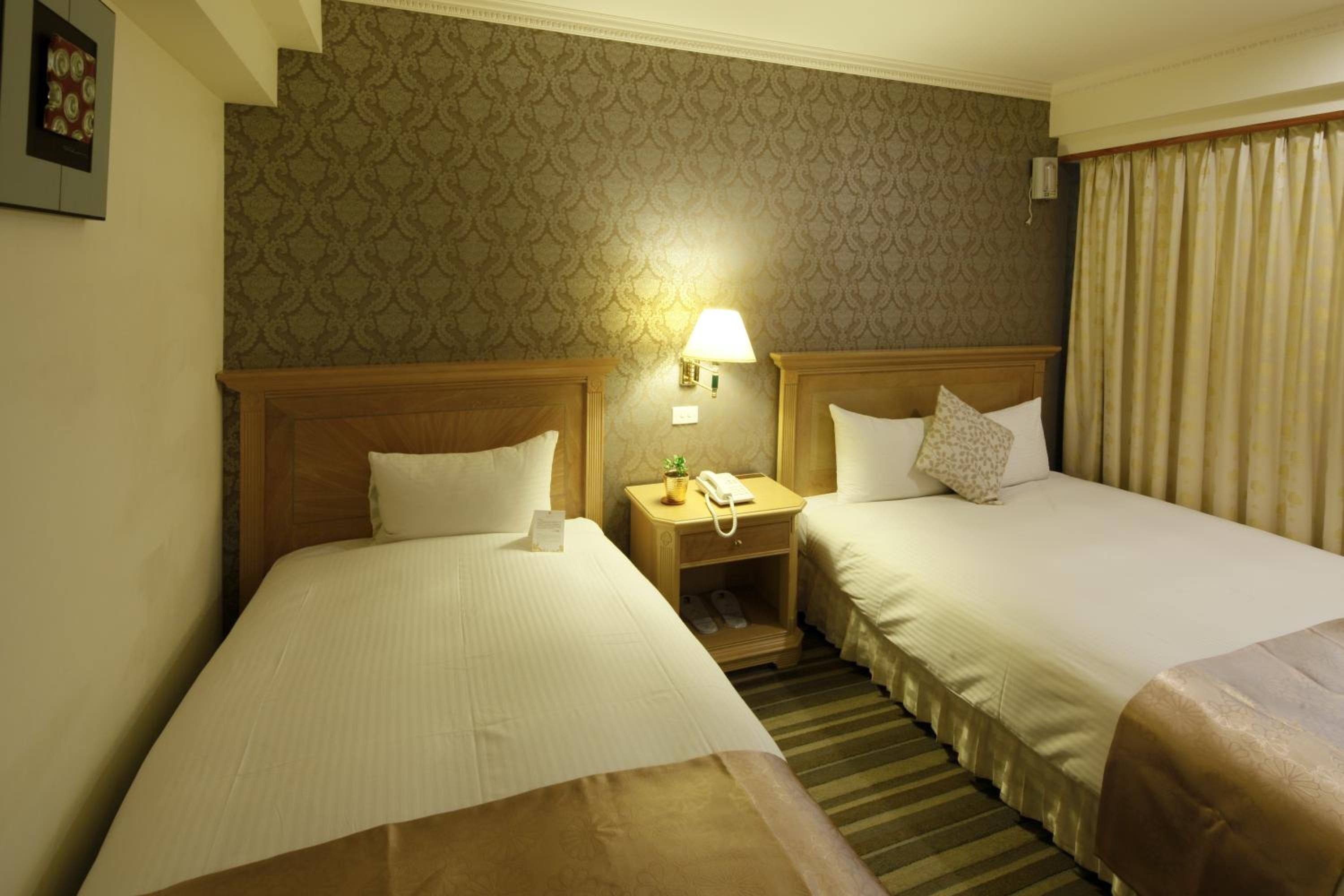 A hotel room with two beds, a nightstand with a lamp, and a small table with a phone and a plant.