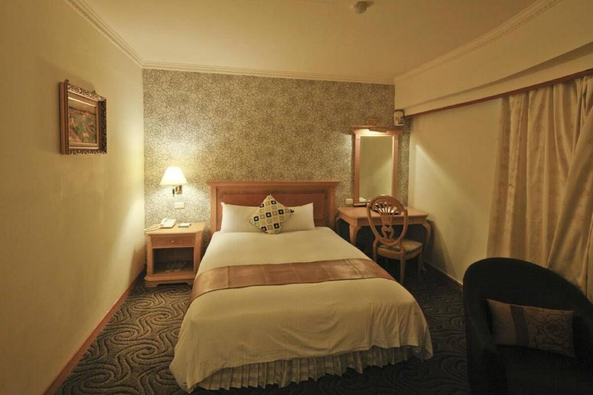 A hotel room with a bed, bedside table, lamp, desk, chair, and a large mirror.