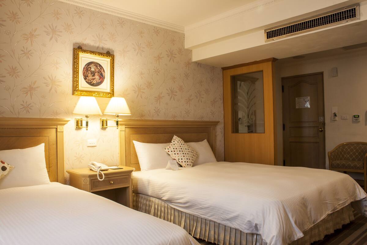A hotel room with two beds, a nightstand with a phone, wall-mounted lamps, and a framed picture on the wall.