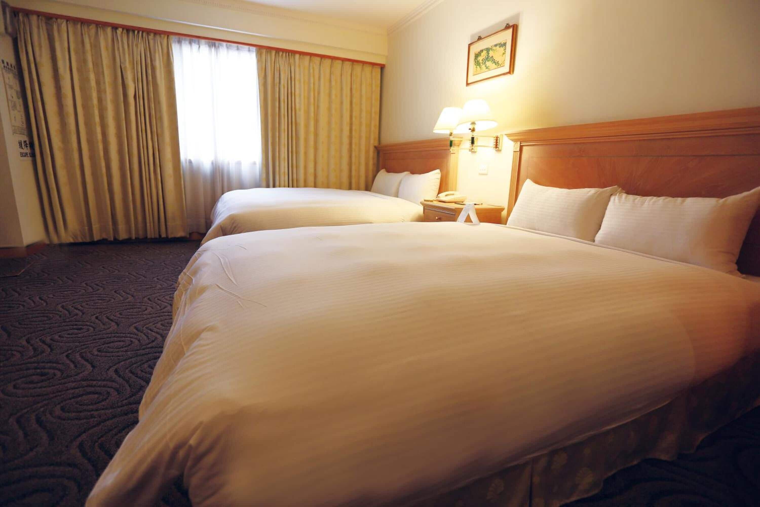 A hotel room with two beds, a wooden headboard, a nightstand with a lamp, and a large window with curtains.