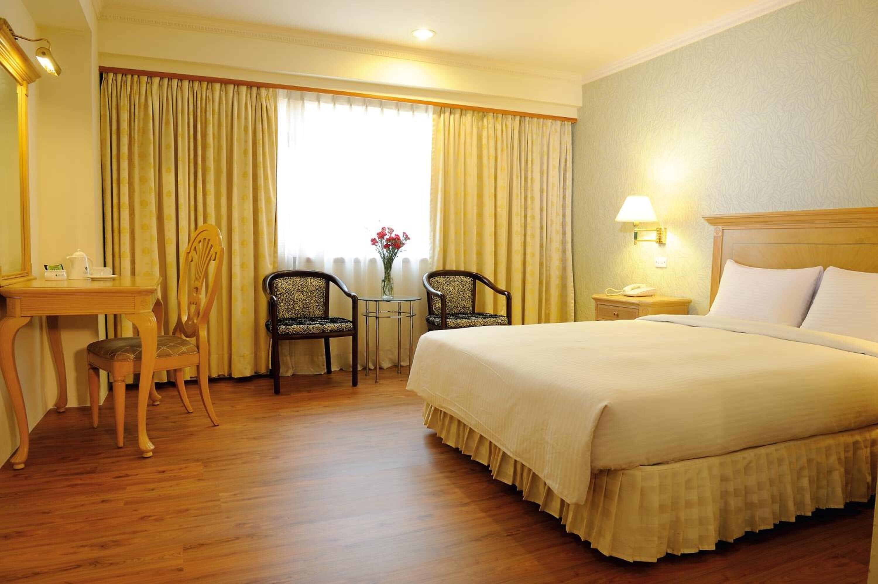 A hotel room with a large bed, a desk with a chair, a small table with a chair, and a window with curtains.