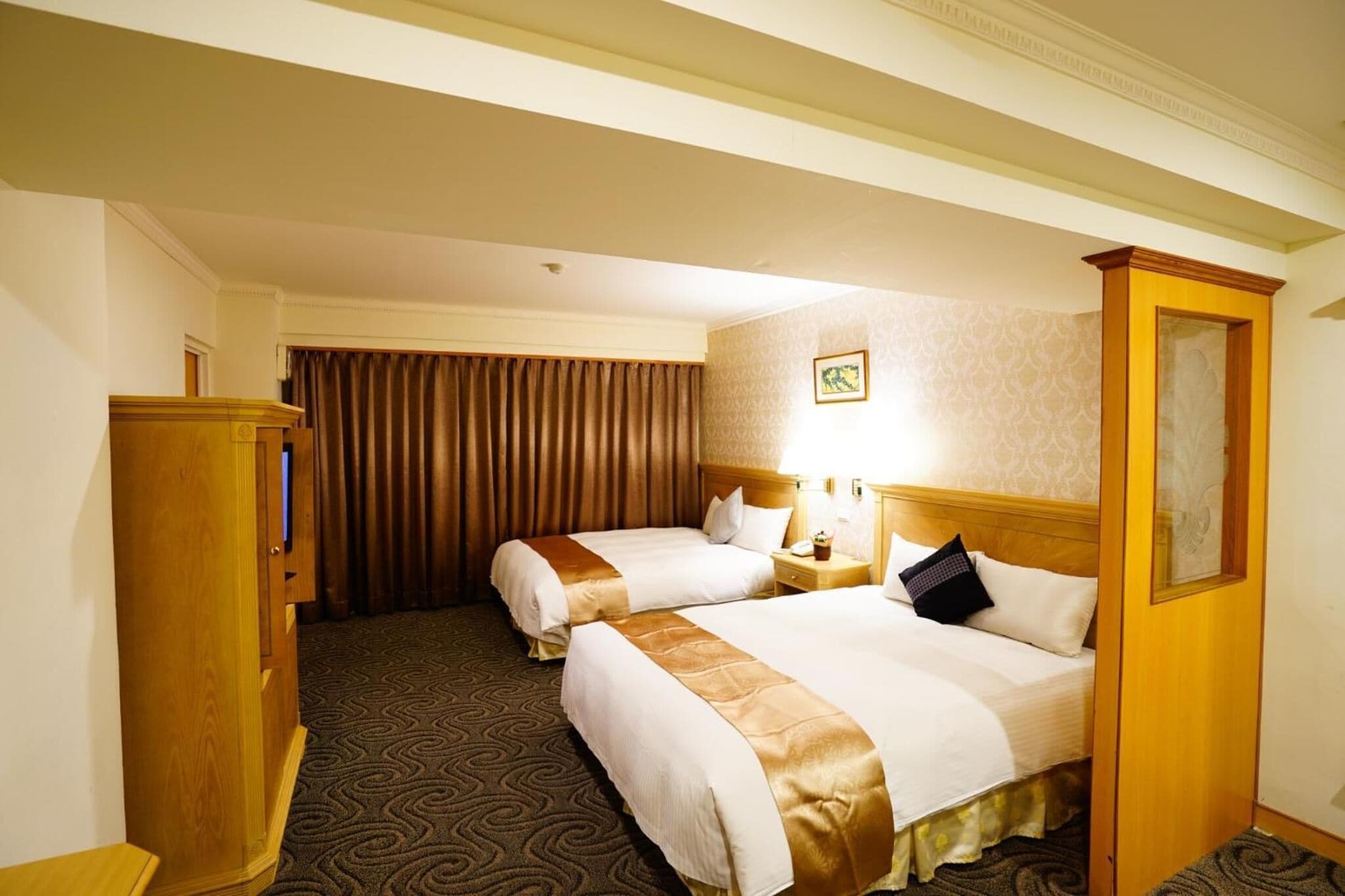 A hotel room with two beds, a wooden wardrobe, a nightstand with a lamp, and a patterned carpet.
