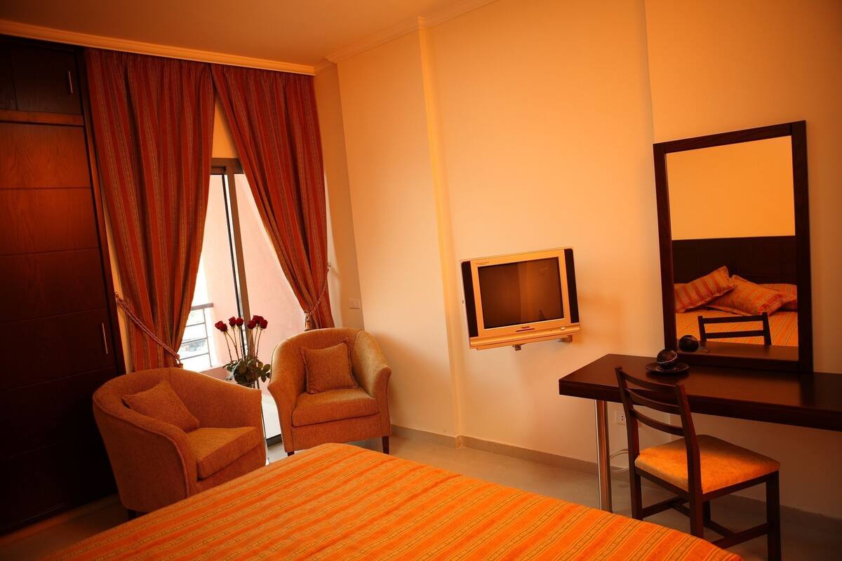 A hotel room with a bed, two armchairs, a desk with a chair, a television, and a mirror.