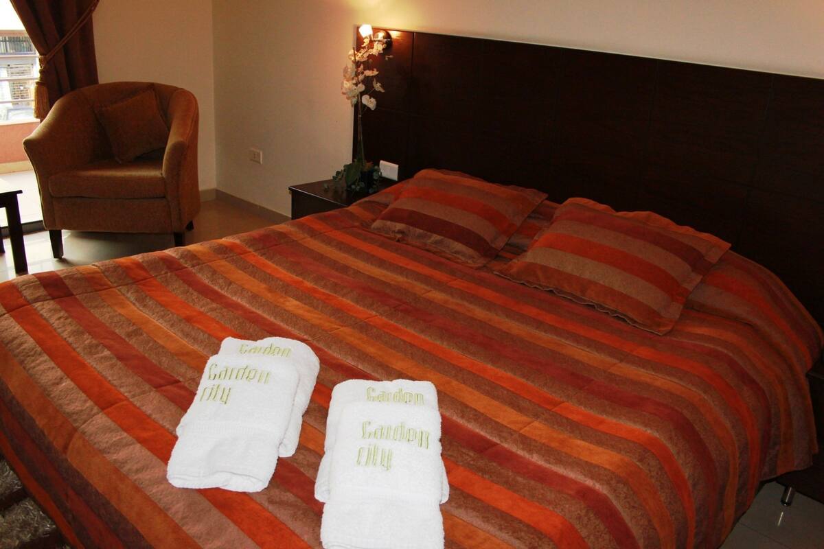 A hotel room with a large bed, two pillows, a nightstand with a lamp, a chair, and two towels with "Garden City" written on them.