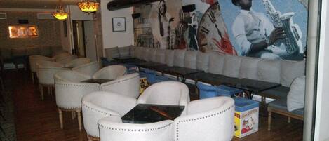 Un restaurante con una larga mesa compartida, sillas blancas tapizadas y un mural de un saxofonista.