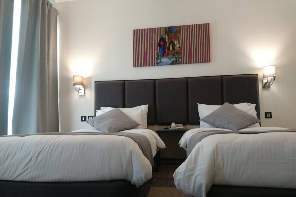 Ein Hotelzimmer mit zwei Betten, einem Kopfteil, Nachttischlampen, einem Gemälde und einem Fenster mit Vorhängen.