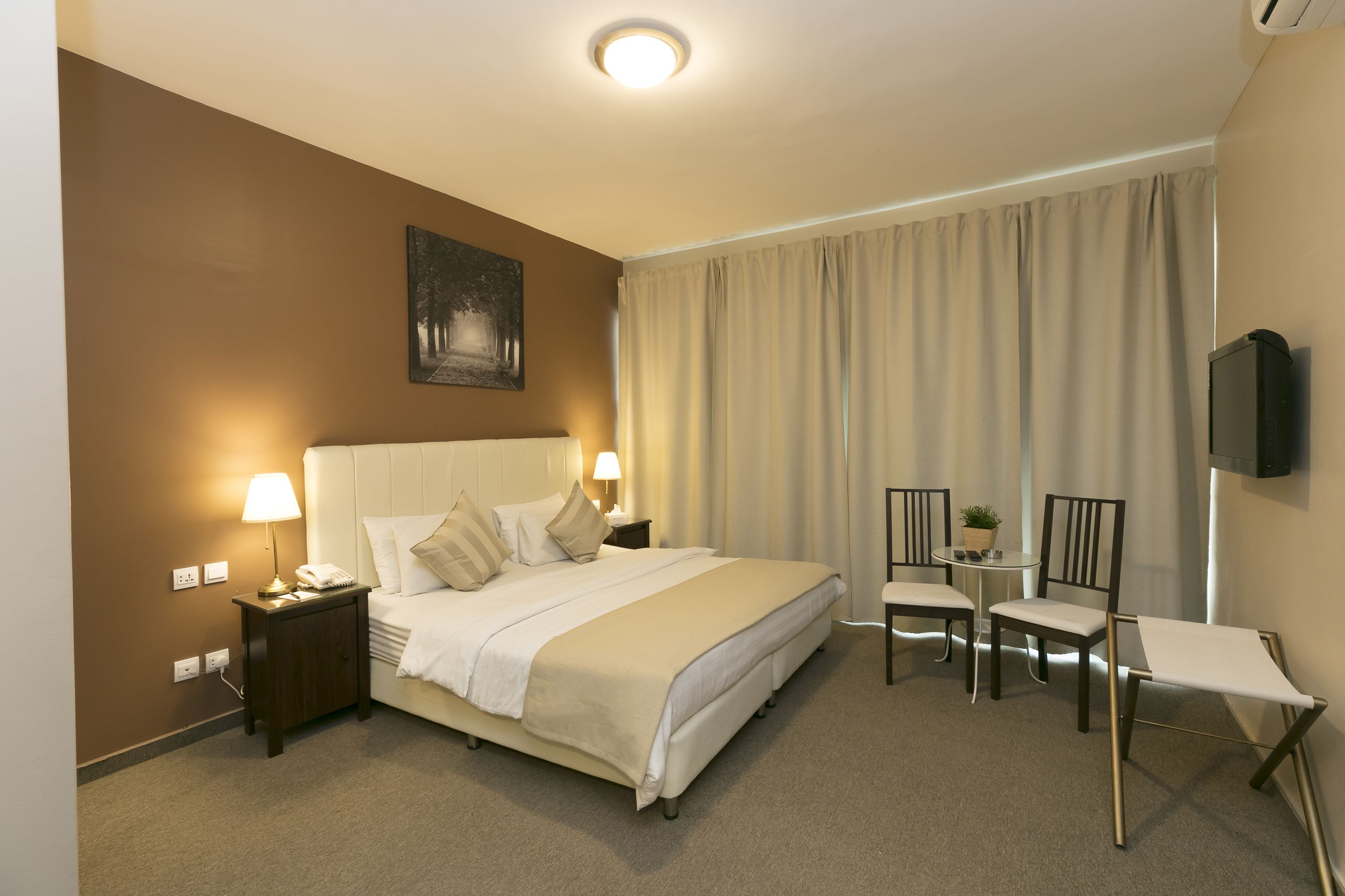 A hotel room with a bed, bedside lamps, a small table with chairs, a TV, and a painting on the wall.