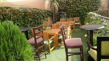 Un espacio al aire libre con mesas y sillas de madera, rodeado de follaje y un seto.