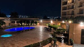 A nighttime outdoor pool area with a circular pool, lounge chairs, and tables, surrounded by greenery and buildings.