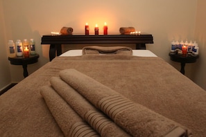 A neatly made bed with rolled towels, a headboard, and a bedside table with candles and toiletries.