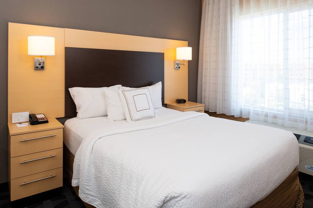 Una habitación de hotel con una cama grande, dos mesitas de noche, un tablero auxiliar, un aparador y un ventanal con cortinas traslúcidas.