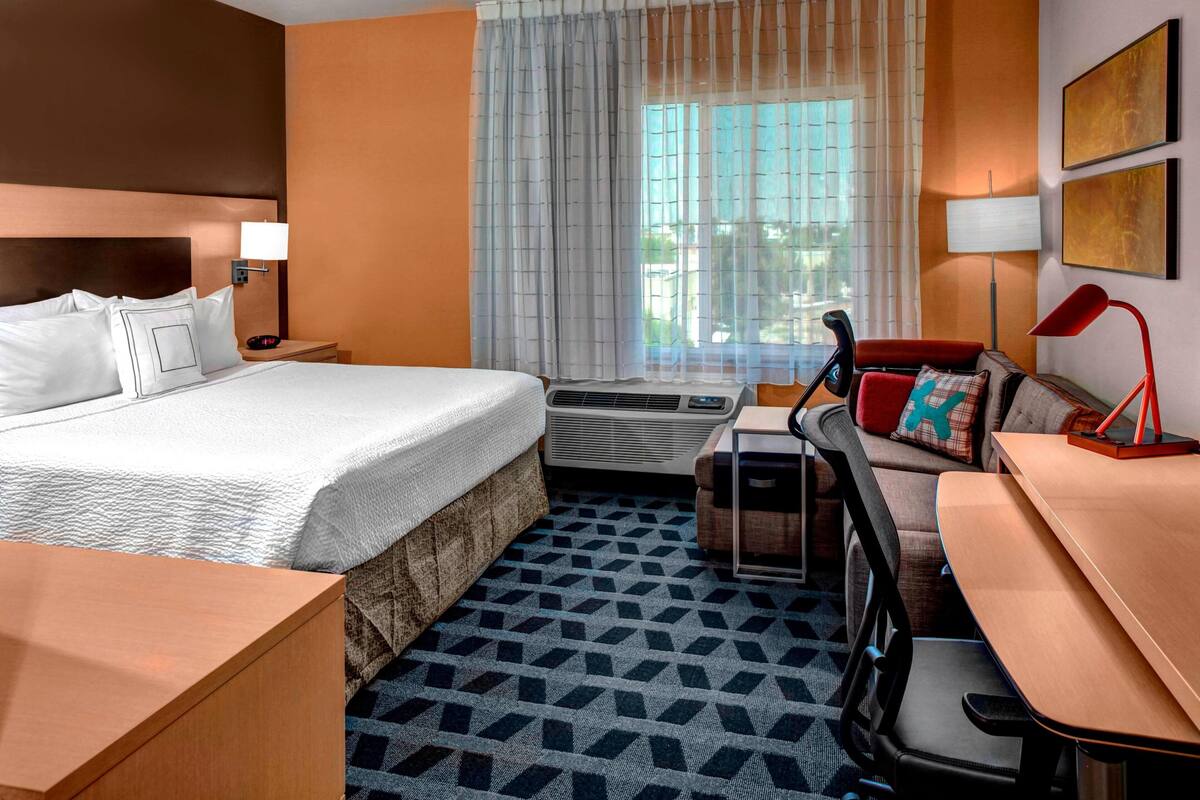 Habitación de hotel con cama, escritorio, silla, sofá, lámpara y ventana con cortinas.